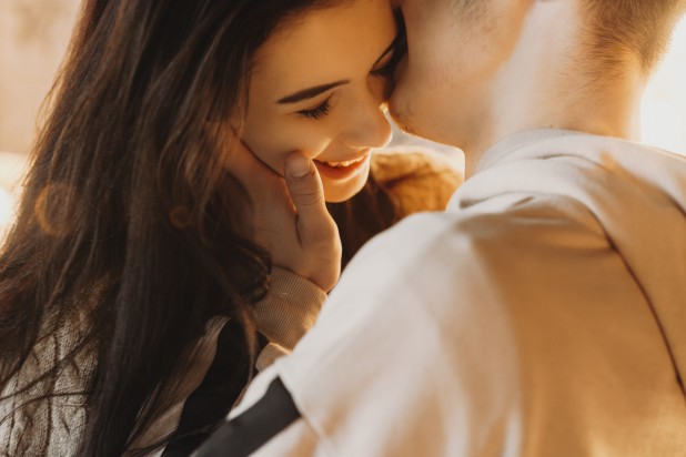 Beautiful close up portrait of an caucasian young couple embracing against light, while boy is kissing his girlfriend cheek which is smiling. (Beautiful close up portrait of an caucasian young couple embracing against light, while boy is kissing his g